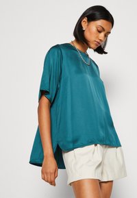 Teal satin top med korte ærmer og en afslappet pasform, med en rundet nederkant og diskrete sideslids. Sammen med cremede shorts.