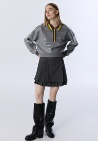 Grijze paillettentrui met een meerkleurige kraag, gecombineerd met een geplooide grijze rok en hoge zwarte leren laarzen.