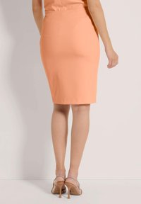 Pfirsichfarbene Bleistiftrock mit glatter Textur und taillierter Form, kombiniert mit transparenten High Heels, die einen Glitzereffekt aufweisen.