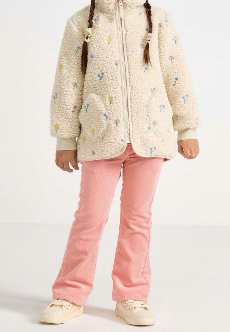 Enfant portant une veste en polaire crème à motif floral, un pantalon en velours côtelé rose et des baskets crème, debout devant un fond blanc uni.
