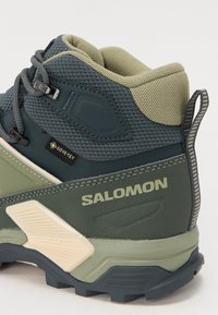 Žali ir tamsiai mėlyni žygio batai su tekstūruotu tinkliniu apavu, GORE-TEX etiketėmis ir kontrastingais kreminiais akcentais ant pado ir logotipo.