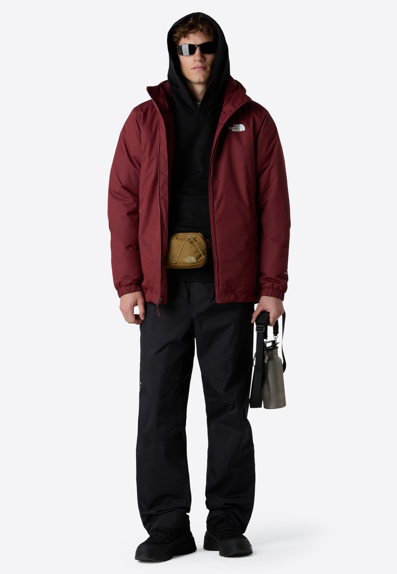 Giacca impermeabile rossa sopra un felpa nera, abbinata a pantaloni neri. Gli accessori includono una borsa a vita beige e una borraccia in acciaio inox.