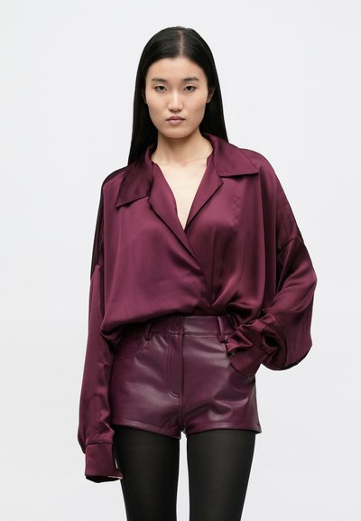 Vino rdeča satenasta srajca z razširjenim ovratnikom in dolgimi, ohlapnimi rokavi, kombinirana s prilegajočimi bordo usnjenimi kratkimi hlačami, nosena s črnimi nogavicami.