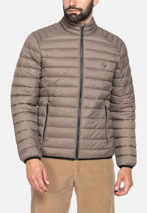 Uomo che indossa una giacca trapuntata taupe con zip nera e tasche laterali, abbinata a pantaloni in velluto a coste marrone chiaro.