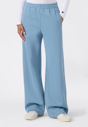 Persona che indossa pantaloni della tuta larghi azzurri chiaro con vita elastica e sneakers bianche, in piedi con una mano in tasca.