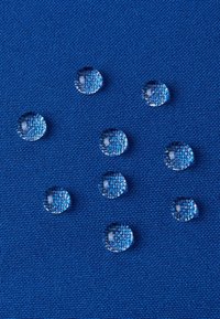 Klare Wassertropfen auf einer strukturierten blauen Stoffoberfläche, mit runder Form und reflektierender Qualität, die in der Größe variieren und gleichmäßig verteilt sind.