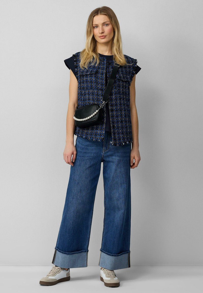 Blauwe tweed vest met franje randen over een donker shirt, gecombineerd met een brede denim broek en een kleine zwarte tas met een parelstrap.
