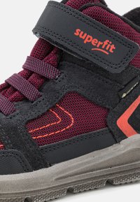 Gyermekcipő fekete és lila hálós felsőrésszel, narancssárga díszítéssel, tépőzáras pánttal és textúrált szürke talppal. Vízálló GORE-TEX anyag.