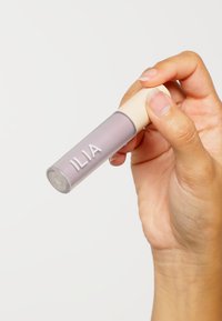 Rund, klar tub med en ljus lavendelfärgad produkt och ett beige lock, märkt "ILIA" i vit text. Hand som håller tuben.