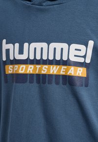 Blauer Kapuzenpullover aus Baumwolle mit einem markanten weißen und orangefarbenen "hummel SPORTSWEAR"-Logo, mit gepunkteten Mustern im Hintergrund.
