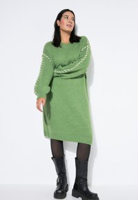 Grünes Strickkleid mit übergroßen Ärmeln und weißen Details an den Armen. Getragen mit schwarzen knielangen Stiefeln und schwarzen Strumpfhosen.