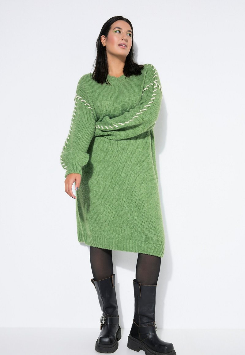 Grünes Strickkleid mit übergroßen Ärmeln und weißen Details an den Armen. Getragen mit schwarzen knielangen Stiefeln und schwarzen Strumpfhosen.