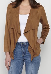 Femme portant une veste marron ouverte sur un haut blanc et un jean bleu, avec une main dans la poche de la veste.