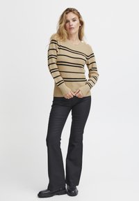 Pull en ribbec beige avec des rayures horizontales noires, manches longues avec des bords côtes, et détails de boutons aux épaules. Associé à un pantalon noir évasé.