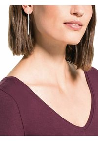 Silverförlängda hoopsörhängen som bärs av en modell med mediumlång brunt hår, mot en bakgrund som visar en djup V-ringad bourgogneblus.