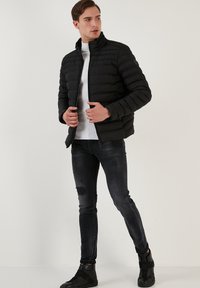 Geacă puffer neagră cu guler înalt, design quilted, închidere cu fermoar și două buzunare laterale, purtată peste o cămașă albă și blugi negri cu aspect uzat.