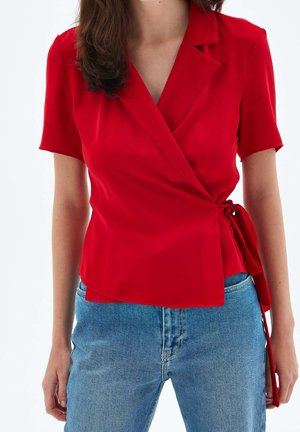 Femme portant un chemisier rouge à manches courtes, noué sur le côté, associé à un jean denim bleu taille haute sur un fond uni.