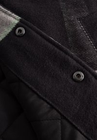 Černá tkanina z vlněné směsi s prošívanou černou podšívkou. Obsahuje velké knoflíky, hladký povrch a jemný zelenošedý vzor.