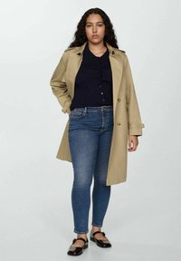 Bež trench kaput preko tamnog dukserice, uparen s plavim uskim trapericama i smeškim loafericama. Neutralna pozadina bez izraženih uzoraka.