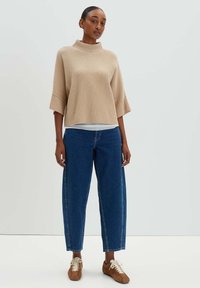 Beiger Ripppullover mit Stehkragen und weiten Ärmeln, kombiniert mit blauen hochgeschnittenen gerade geschnittenen Jeans und braunen Schnürschuhen.