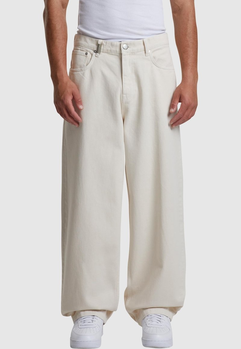 Homme portant un pantalon ample beige clair, des baskets blanches et un haut blanc sans manches sur fond uni.