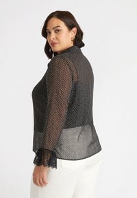 Blusa nera a pois con tessuto trasparente, colletto e polsini in pizzo. Presenta una sezione posteriore solida a contrasto e una vestibilità comoda.