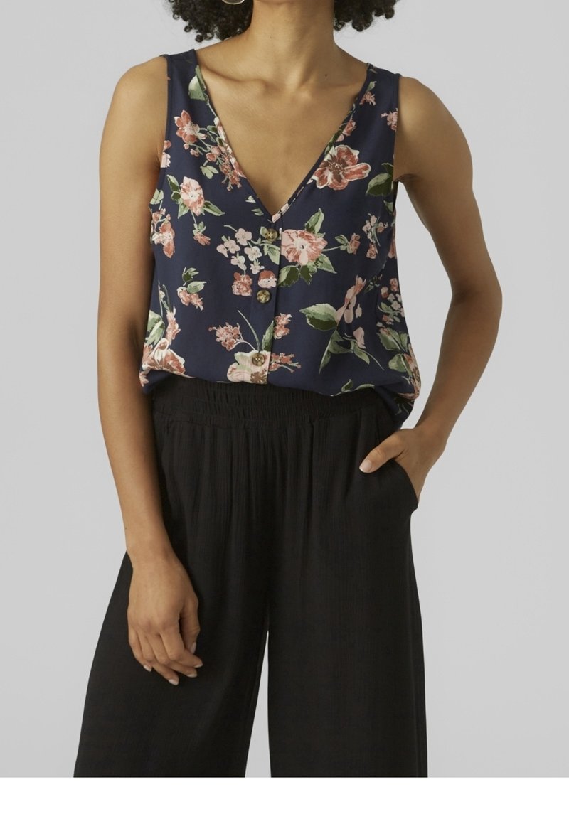 Hedvábný top na ramínka s květinovým vzorem v námořnické modři, s hlubokým V výstřihem, malými knoflíky a bez rukávů. Kombinován s černými, texturovanými kalhotami.