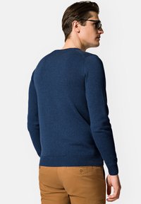 Granatowy sweter z dzianiny z długimi rękawami, okrągłym dekoltem i prążkowanymi mankietami; zestawiony z beżowymi chinosami z ozdobnym guzikem z tyłu.