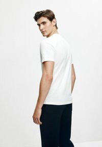 Biała bawełniana koszulka z krótkim rękawem i klasycznym okrągłym dekoltem, noszona z ciemnymi spodniami, ukazująca gładką fakturę i dopasowany krój.