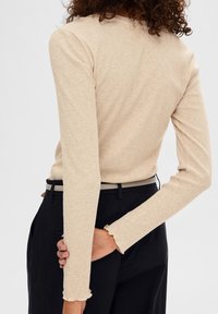 Top beige a costine con maniche lunghe e scollatura rotonda, con polsini arricciati, abbinato a pantaloni scuri a vita alta e una cintura sottile.