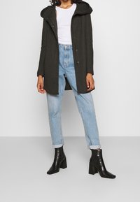 Abrigo verde oscuro con capucha y cierre de botones, llevado sobre una blusa blanca. Combinado con jeans de corte relajado en azul claro y botines negros.
