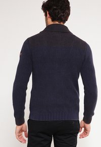 Ciemnoniebieski sweter z dzianiny z fakturowanym wzorem, z prążkowanym kołnierzem i mankietami. Posiada kontrastującą górną część oraz subtelny detal z logo.