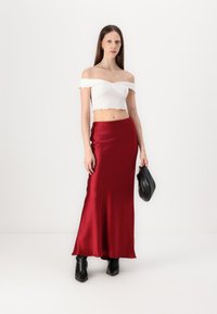 Biely crop top bez ramienok s volánovými okrajmi, skombinovaný s červenou saténovou maxi sukňou. Modelka drží čiernu kabelku a má na sebe čierne zdravotné topánky.