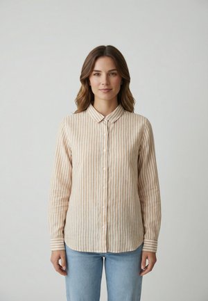 Frau mit schulterlangen braunen Haaren, die eine beige-weiße vertikal gestreifte Knopfleiste und hellblaue Jeans trägt, steht vor einem einfarbigen Hintergrund.