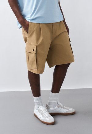 Άνδρας που φοράει μπεζ cargo σορτς, ανοιχτό μπλε πουκάμισο, λευκές κάλτσες και λευκά αθλητικά παπούτσια με καφέ σόλες, όρθιος σε γκρίζο πάτωμα.