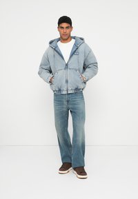 Chaqueta de mezclilla con capucha en azul claro, con cierre de cremallera, bolsillos frontales y puños elásticos, combinada con jeans azules claros y zapatillas marrones.