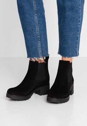 Bottines en daim noir avec des panneaux latéraux élastiques, un talon épais et une semelle en caoutchouc, associées à un jean bleu court avec des ourlets effilochés.