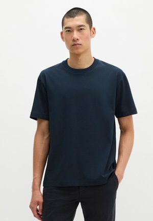 Jonge man met kort haar die een effen donker marineblauw T-shirt met korte mouwen en een zwarte broek draagt, staat met één hand in zijn zak.