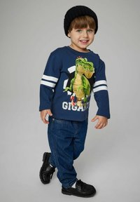 Maglietta a maniche lunghe blu navy con grafica di un dinosauro verde e testo "GIGANTIC", dettagli a righe bianche. Abbinata a pantaloni in denim blu e scarpe nere.