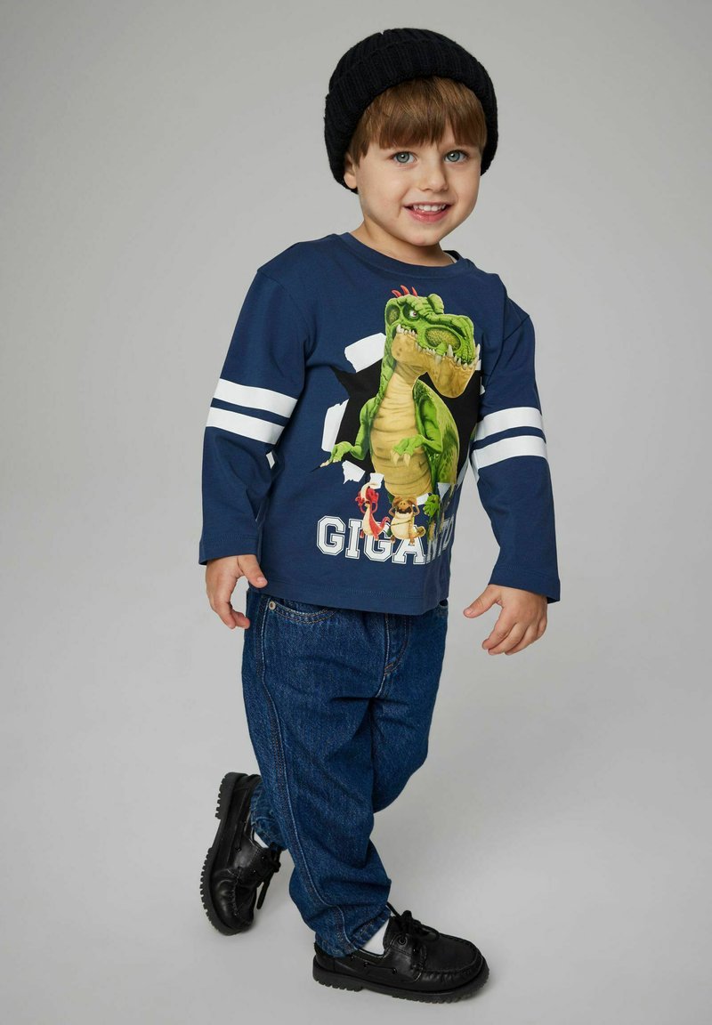 Maglietta a maniche lunghe blu navy con grafica di un dinosauro verde e testo "GIGANTIC", dettagli a righe bianche. Abbinata a pantaloni in denim blu e scarpe nere.