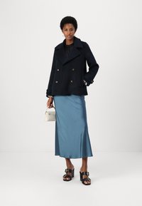 Marine dobbeltspent peacoat, sateng teal glideskjørt, svarte blokkhelte sandaler med gullakenter, og en liten hvit håndveske.