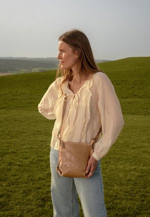 Vrouw met lang haar die een licht gerimpeld blouse, spijkerbroek en een beige crossbodytas draagt, staand op met gras begroeide heuvels bij zonsondergang.
