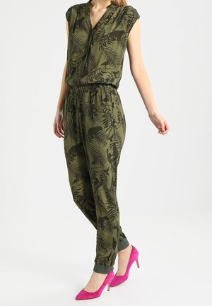 Kvinde iført en olivengrøn jumpsuit med sorte blad- og dyreprint kombineret med lyserøde højhælede sko, stående mod en ensfarvet baggrund.