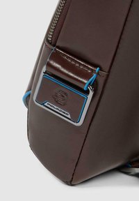 Esquina de bolso de cuero marrón oscuro con cremallera visible y placa metálica con logo que presenta detalles en azul y emblema de marca en relieve.