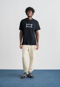 Μαύρο μπλουζάκι με μεγάλο γκρι γραφικό σχέδιο, λεπτομέρειες στο πλάι, συνδυασμένο με κρεμ παντελόνι και μαύρα αθλητικά παπούτσια, σε μπλε φόντο.