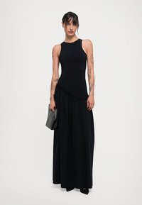 Mustaton olkaimeton mekko, jossa on istuva bodice ja levenevä maksihame. Sujuva kangas, sivusauman yksityiskohta. Malli pitelee teksturoitua mustaa clutchia.