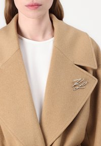 Cappotto in lana di cammello con ampi revers e spilla argentata. Il tessuto appare morbido e textured. Sotto, è visibile una maglietta bianca.