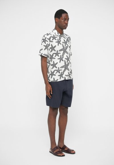 Majica s kratkimi rokavi z vzorcem črnih morskih zvezd na belem ozadju, v kombinaciji s temnomodrimi kratkimi hlačami in rjavimi sandali, oseba obrnjena levo.