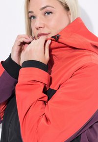 Giacca impermeabile rossa e nera con cappuccio, dotata di colletto con zip, cordini regolabili e accenti viola a contrasto.