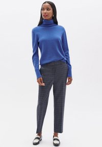 Un pull col roulé bleu en tissu doux associé à un pantalon en tissu à carreaux gris. Le mannequin porte des chaussures noir et blanc, mettant en valeur une coupe ajustée.